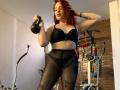 ElizabethDom with red hair, black bra, and sheer black pantyhose, holding a kettle, standing, showing tattoos on thighs, looking down, in a room with gym equipment.
