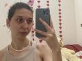 Sanella with light skin and brown hair in a messy bun, wearing a beige tank top and white bead necklace, holding a gray phone, showing her face and small tits.