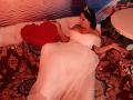 AmberSunshine con il seno scoperto, vestita con un abito bianco a spalle scoperte, sdraiata su un divano a motivi rossi e marroni, che tiene un cuscino a forma di cuore rosso, indossando una tiara, in una stanza illuminata di rosso..