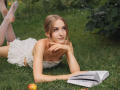 EvaDaly with blonde hair, lying on grass, wearing a white dress, legs up, reading a book, apple near, smiling, hands under chin, bare tits, green background.