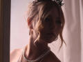 RosaVance has blonde, wavy hair in a messy updo with black floral hairpiece. She's wearing pearl earrings and a double pearl necklace, looking slightly to the side.