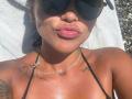 MajoJones with tan skin, full pink lips, black bikini top, tattoos on chest and shoulders, wearing dark sunglasses, lying on a white towel, sunlight on her face.