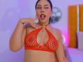 MarilynHills with tan skin, black hair, and red lipstick, wearing a red fishnet bralette, sticking her tongue out and touching her lips with her finger. Boobs, tongue out.
