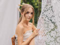 EvaDaly with small tits, wearing a white off-shoulder top, holding lace curtain, sitting outdoors, wooden chair, blonde hair in ponytail, subtle smile, green background, daylight.