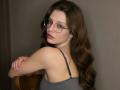 StaceyAverbach with long brown hair, glasses, and a gray tank top, showing her bare shoulders and arms, sitting on a wooden chair, looking back with a slight smile.