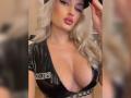 MademoiselleBella has big, shiny black latex tits with a deep cleavage, wearing a black cap, and has platinum blonde wavy hair, pink lips, and a sultry expression.
