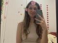 Sanella with long brown hair, pink flower headpiece, beige top, holding green phone, smiling, white wall, red heart decals, beige teddy bear, pearl necklace, cute, mirror selfie.