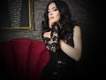 MistressTaylor with black lace dress, curly black hair, and pink lipstick, touching her chin with her right hand, red couch in background. Her tits are visible through lace.