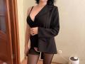 EmmaNoa indossa un blazer nero, un reggiseno nero e collant neri trasparenti, mostrando i suoi seni attraverso un'apertura. La sua mano è sul ventre, ha i capelli neri e il viso non è visibile..