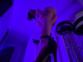 SherylHill with black high heels, tan skin, tattoo on right side, and long dreadlocks, standing in purple-lit room, arms behind head, looking down at camera.