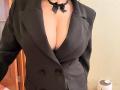 EmmaNoa indossa un blazer nero con una profonda scollatura a V, che mette in mostra un seno generoso, un collare nero con un fiocco, bottoni doppi e una giacca leggermente aperta..