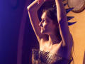 NikaFlorence with arms raised, wearing a spiked headpiece and mesh top, showing tits and a profile view, in warm, purple-lit light. Her face is slightly turned to the side.