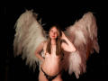 AmberWoody with white angel wings, topless, wearing black lace panties, hand in hair, smiling, pink lipstick, light skin, brown hair, small tits, slightly pregnant, dark background.