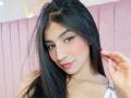 NaiaSanders has long black hair, a nose ring, and pink blush. She's smiling with winged eyeliner, pink lips, and a white top. She's touching her hair with a hand.