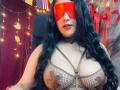 CristalZafiro with long black hair, wearing a red blindfold, brown fishnet bra with black pasties, and a black choker, sitting in a chair, showing her tits.