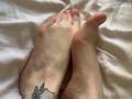 Mettis's bare feet with a small black and gray tattoo on the right ankle, toes slightly spread, resting on a white bedsheet. Visible veins and slightly pink soles.