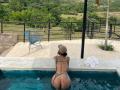 AlessiaFerry with tattoos, butt in a blue leopard-print bikini, hat, in a pool, facing away, white towel on stone edge, black metal fence, blue chair in background.