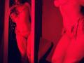 Red-lit photo of Tisia in a sheer lingerie set with chain details, showing her tits, ass, and pussy in a mirror, standing with one hand near her mouth.