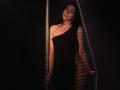 TriciaBarnes with long black hair, wearing a black one-shoulder dress, standing in a dark, striped-lit area, hands on a metal frame, exposing her ass.