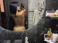 MajoJones, naked with black hair, facing a mirror, showing a tattoo on her back and her bare ass, holding a phone, in a bathroom with black marble and toiletries.