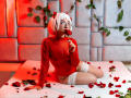 KanelaMiller mit weißem Bob, Brille, rotem Samms-Body, weißen Spitzen-Overknees, die eine Rose an den Mund führt, sitzend auf einem Bett mit Rosenblüten, rote und graue Wand..