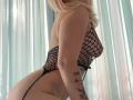 BaeonLive with blonde hair, wearing a black fishnet bodysuit, showing her big ass, small tits, and tattoos on her left arm, standing in front of sheer curtains.