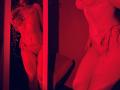 Red-lit photo of Tisia with lace lingerie, showing her toned body, small tits, and trimmed pussy, reflected in a mirror, holding a phone to her face.