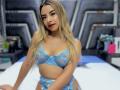 CloeRouss met blond haar, rode lippenstift, gekleed in blauwe kant lingerie, die haar borsten en vulva laat zien, zittend op een bed, kijkend naar de camera, met een glimlach..
