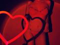 Red-lit photo of Tisia in lace lingerie, showing her tits and pussy, with neon heart and black heart shadows on her skin, standing with hands on her hips.