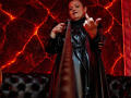 VanesaMoka in a black latex dress, pointing with one finger, holding a long, narrow object, looking confident, with a tufted black leather couch in the foreground.