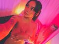 Yurimy with dark hair, looking up, topless, covering her tits with a hand, pink and orange lighting, tattoos on her arm, white nails, bracelet on her wrist.