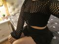 Niferttiti wearing a black fishnet top, exposing small tits, and a black skirt, sitting on a bed with gray heart-patterned bedding. Niferttiti's hands rest on her thighs.