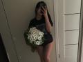 Olieri with medium Asian skin tone, black shirt, holding white daisies, standing, phone in hand, mirror selfie, visible ass, bare thighs, long dark hair, dim room.
