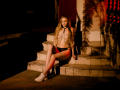 MiaMeowa with long blonde hair, wearing a chain top, black panties, and white sneakers, sitting on stone steps, holding a black clutch, in dim, red-lit surroundings.