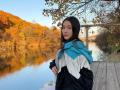 AshleyNaone with long dark hair, wearing a blue and white jacket, sitting on a wooden dock, looking over her shoulder at a calm, reflective river with autumn trees.