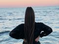 Niferttiti with long brown hair, black lace shirt, hands on hips, standing in front of a blue ocean, sunset in the background.