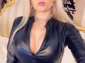 Mademoiselle Bella, com seios grandes, vestindo uma blusa preta brilhante com zíper, batom rosa, um colar com pedras, cabelo loiro, brincos e um colar, tirando uma selfie..