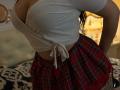 Niferttiti in a white, ribbed, tied-up top showing large, round boobs and wearing a red, plaid, lace-trimmed mini-skirt, standing with hands on hips.