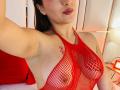 KatarinaNovak in red fishnet lingerie, showing her tits with visible nipples, a small tattoo on her right arm, and a slight smile. Her dark hair is in a ponytail.