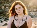 Rubbi with red hair, freckles, black bikini top, tattoos on chest, and multiple necklaces, standing on a sunny beach, sunlight in the background.