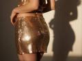 Diantrisy's shiny gold sequined ass, covered in tiny reflective tiles, is illuminated by sunlight, casting a shadow on a plain wall. Her hand rests on her lower back.