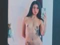Nude LilithDesire with long black hair, small tits, tattoos on her side and lower belly, holding a phone, taking a mirror selfie. Her pussy is partially visible.