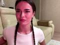 DrusillaMaiava has pink-tinted skin, braided brown hair, and wears a white shirt. She's sitting on a beige leather couch with a small wooden pillow behind her.