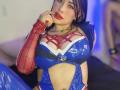 Kkole with big tits, wearing a blue and red latex Spider-Woman costume, finger in mouth, red lipstick, black hair, and a sexy, confident look. Her arm has red webbing.