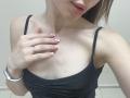 Ramilla's chest with small tits, wearing a black tank top, has a hand with pink nails touching her left breast, showing a ring and a white bracelet.