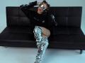 MilagrosEdmisten lounges on a black leather couch, legs spread, wearing a black top with shiny silver sleeves and thigh-high silver boots. She wears reflective sunglasses and red lipstick. Fucking hot.