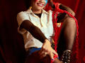 LucieMilner with painted blue face, white shirt, black fishnet sleeves, denim shorts, and red chain, smiling, sitting, showing her ass, wearing fishnet stockings and black boots.