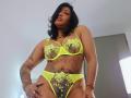Nahirr, wearing neon yellow lingerie with floral lace, has medium tits, a belly button piercing, and tattoos on her arms and stomach, standing with a confident, slightly pouty expression.