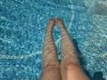 Barbarica's legs are in a clear blue pool with sunlight rippling through the water. Her feet are pointed straight, showing her bare toes. Water reflects light on her skin.