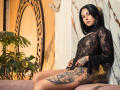 GabyMathew with black lace top, tattooed thighs, and medium tits, sitting on a couch, looking at camera, dark hair, pale skin, marble wall, plant on left.
